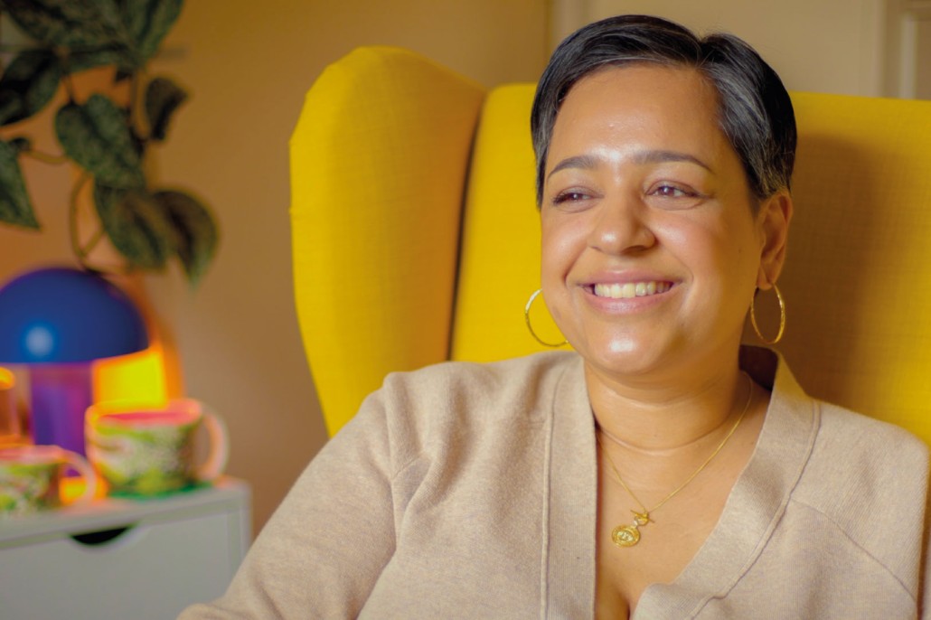 Image of lady sitting on a yellow chair smiling and looking right.